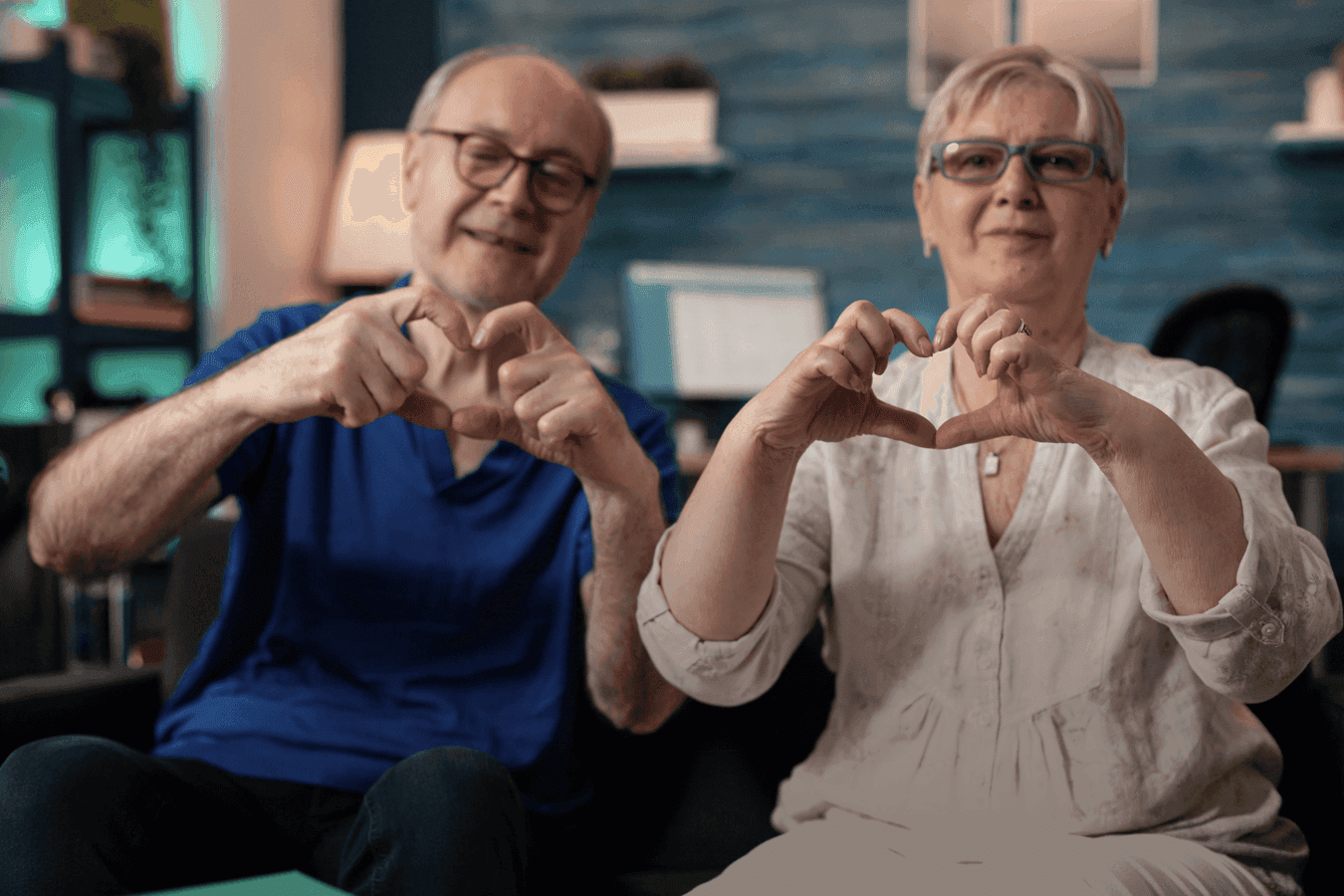 Elderly couple sitting on a couch, smiling and forming heart shapes with their hands in a cozy, warmly lit setting.