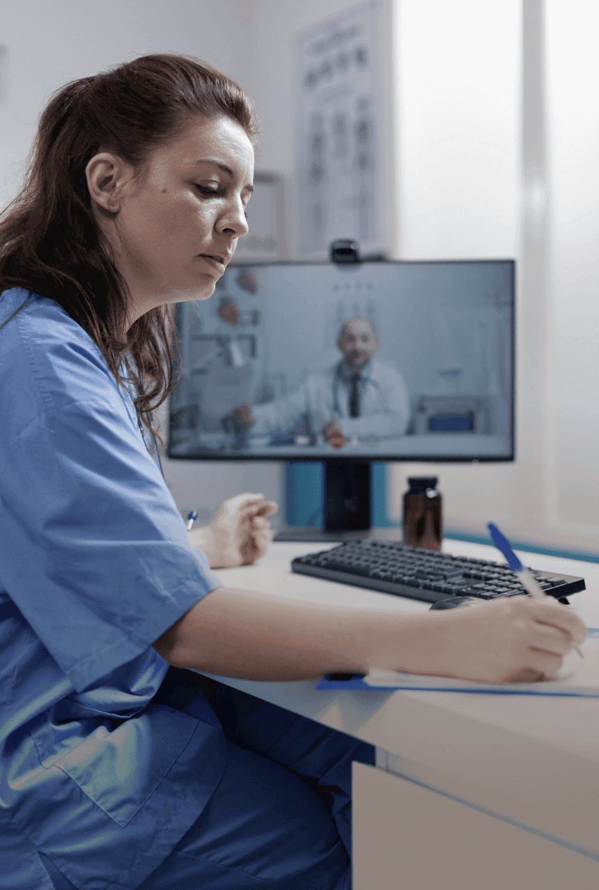 Healthcare professional in blue scrubs taking notes while watching an educational video on a computer in a medical office setting.